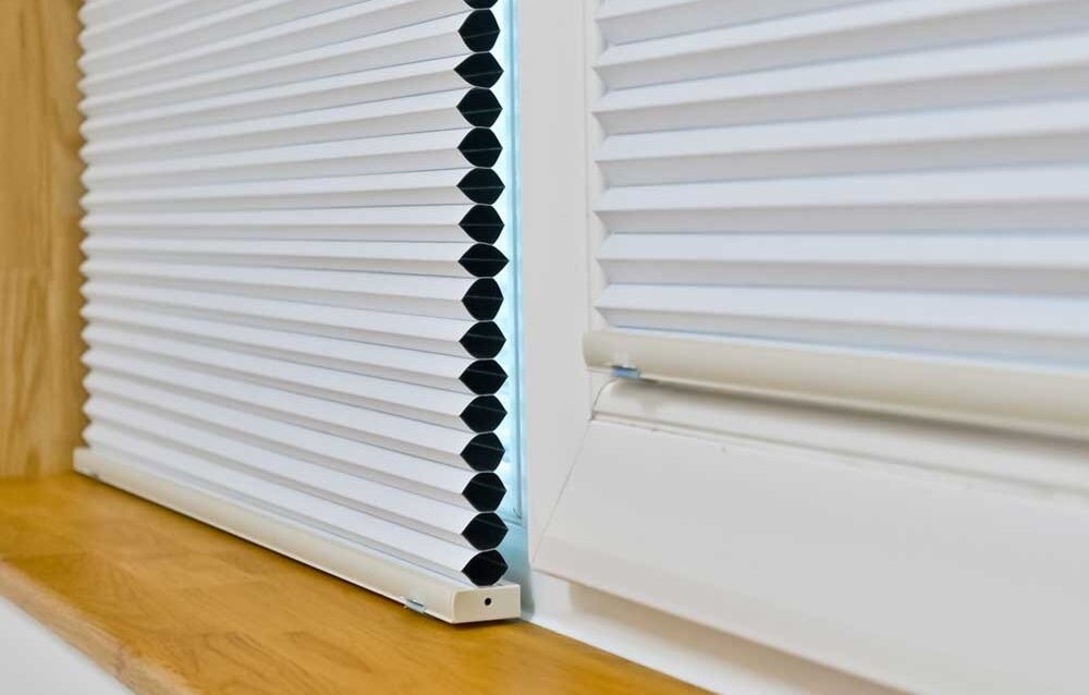 WhatsApp Image 2026-03-18 at 11.17.45 Close-up of a white honeycomb (cellular) window blind partially lowered on a wooden windowsill, showing its pleated structure and side profile.