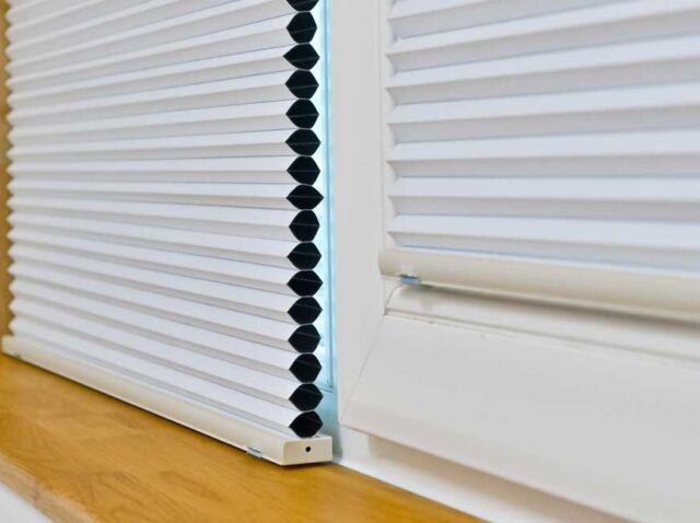 WhatsApp Image 2026-03-18 at 11.17.45 Close-up of a white honeycomb (cellular) window blind partially lowered on a wooden windowsill, showing its pleated structure and side profile.